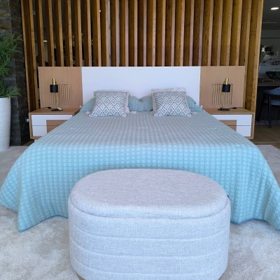 Quarto com cama de casal, roupa azul e almofadas cinza, com puff cinza diante da cama