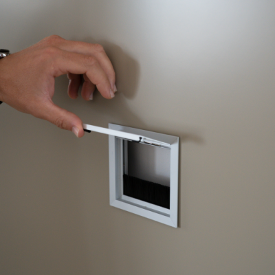 Mão a abrir orifício quadrado numa parede bege com moldura branca e cerdas pretas