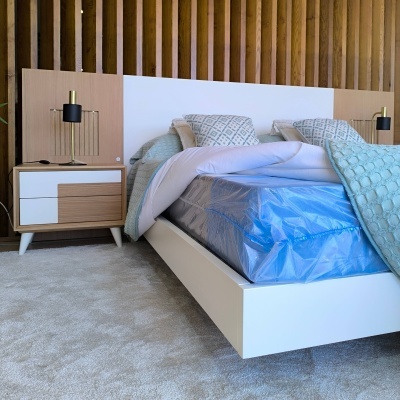 Cama branca de madeira com colchão plástico azul, cabeceira de madeira e branco, roupa de cama azul e cinza, mesa de cabeceira com gavetas, tapete cinzento