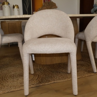 Cadeira estofada bege claro com textura, pernas arredondadas, etiqueta CESARI, sobre carpete de fibra natural, com mesa de madeira ao fundo.