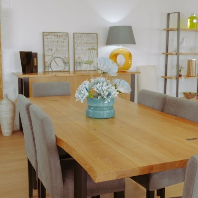 Mesa de madeira com seis cadeiras cinzentas, vaso azul com flores brancas ao centro, decoração e prateleiras ao fundo