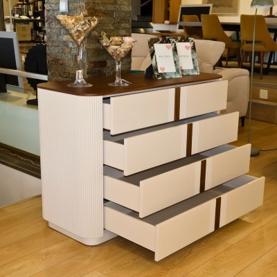 Cómoda branca fosca com detalhes em madeira castanha e cinco gavetas abertas, decorada com recipientes de vidro e quadros, em loja de móveis