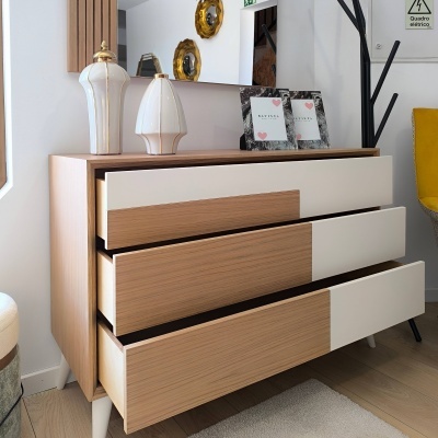 Cómoda em madeira clara com gavetas brancas abertas, candeeiros e quadros em cima, parede com espelho e decoração