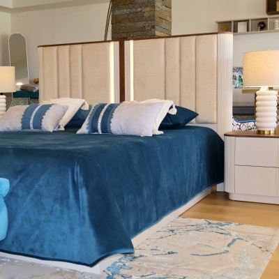 Cama estofada bege com cobertor azul e almofadas, mesa de cabeceira com candeeiro branco em quarto elegante