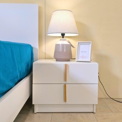 Mesa de cabeceira branca com candeeiro e moldura com texto ao lado de cama com lençol azul