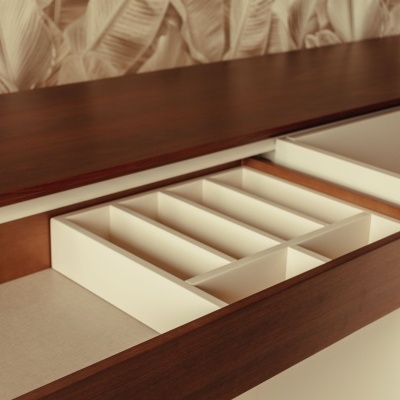 Gaveta de móvel com divisórias internas brancas e acabamento em madeira castanha com papel de parede de folhas bege ao fundo