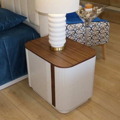 Mesa de cabeceira branca com tampo de madeira e candeeiro branco ao lado de cama azul e cadeira estampada