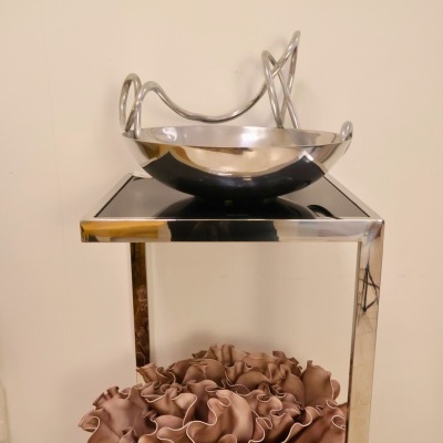 Fruteira metálica curva sobre mesa metálica com almofada castanha pregueada ao fundo