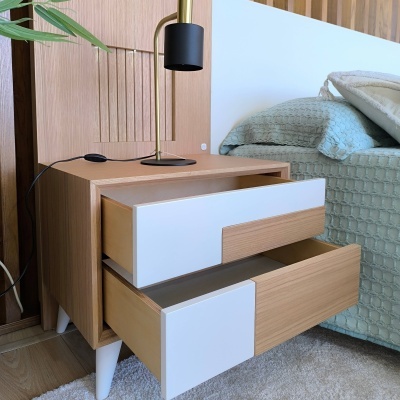 Mesa de cabeceira madeira clara com gavetões brancos ao lado de cama azul e candeeiro dourado