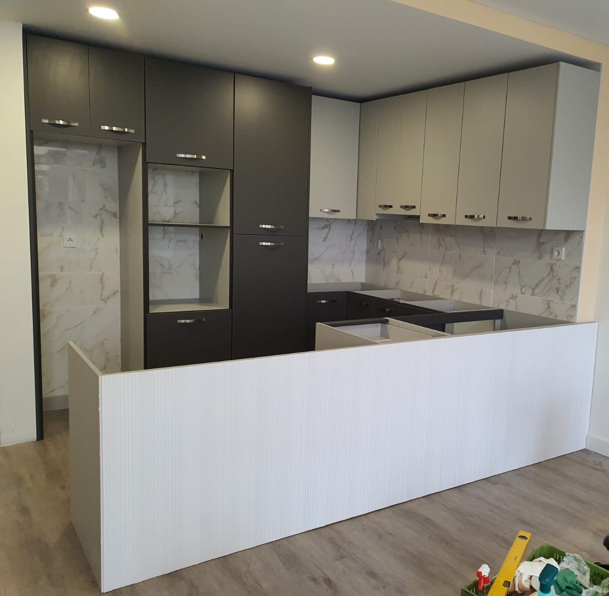 Cozinha moderna com armários cinza escuro e claro, azulejo efeito mármore e bancada branca em fase de montagem.