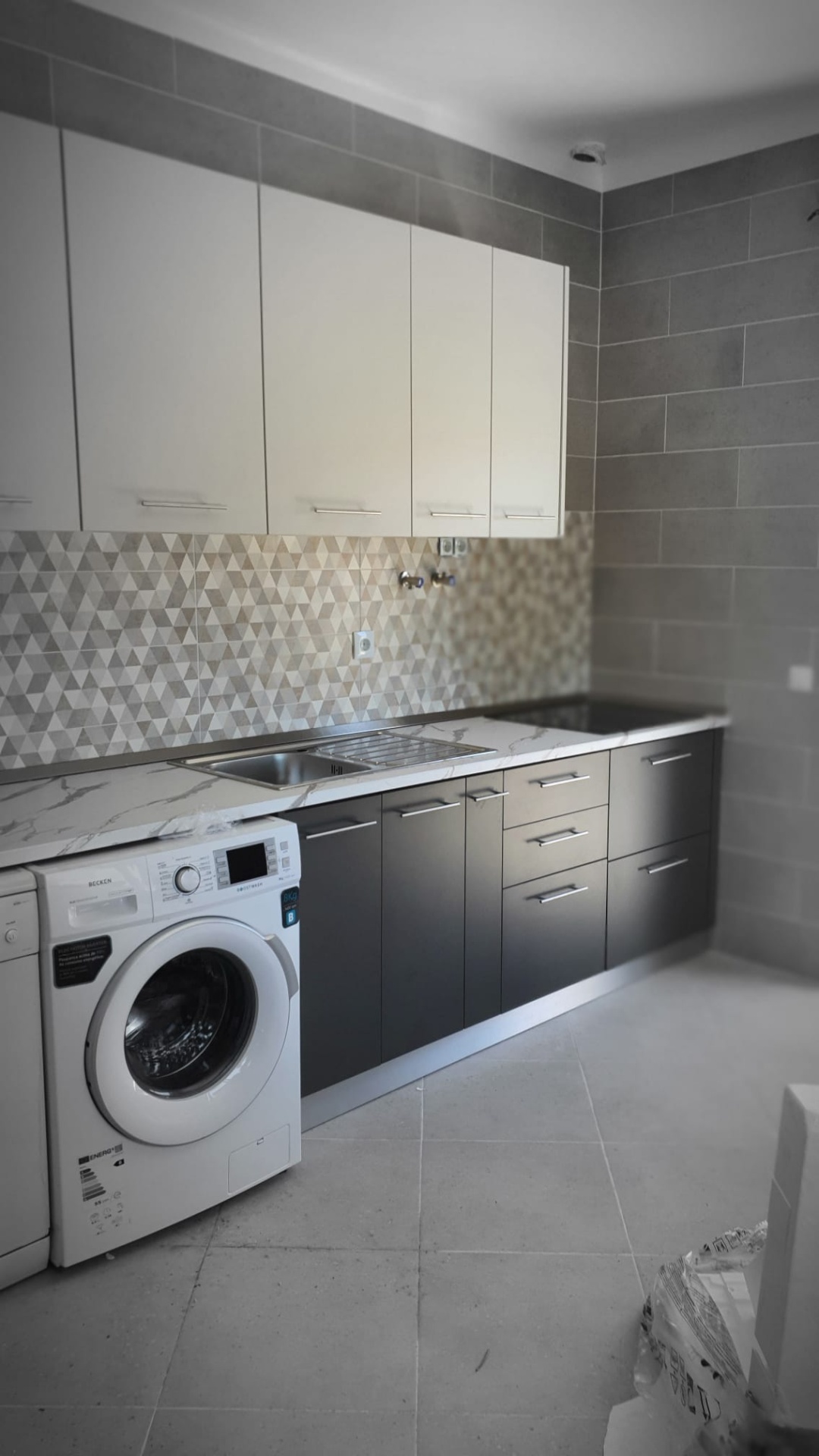 Cozinha moderna com armários brancos e cinza, bancada mármore, lava-loiça inox, máquina de lavar roupa branca e azulejos geométricos.