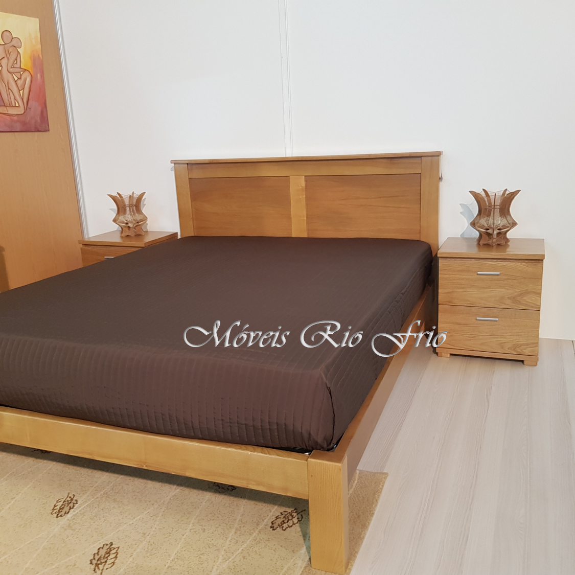 Cama de madeira clara com cabeceira, colchão com lençol preto, duas mesas de cabeceira com gavetas e lâmpadas decorativas