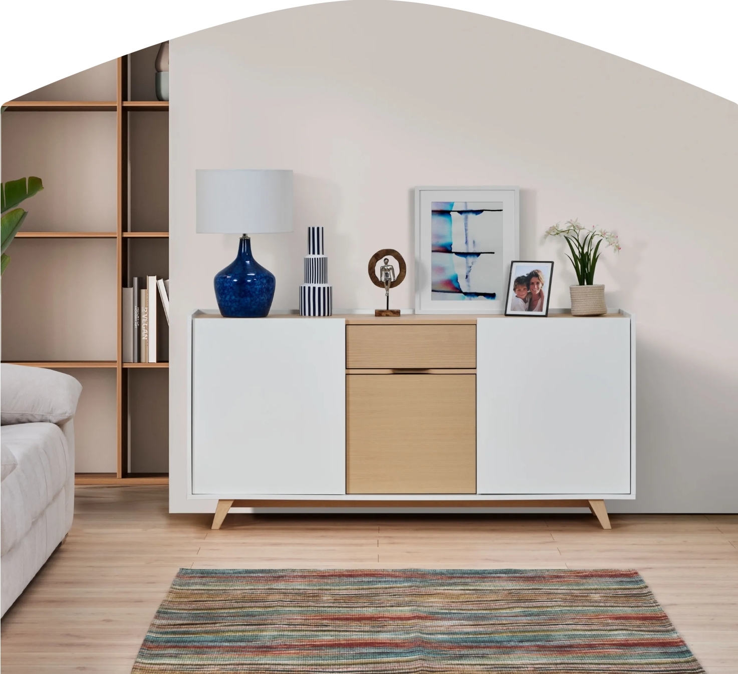 Aparador branco com portas e gaveta em madeira clara decorado com objetos e quadro numa sala com estante e sofá claro
