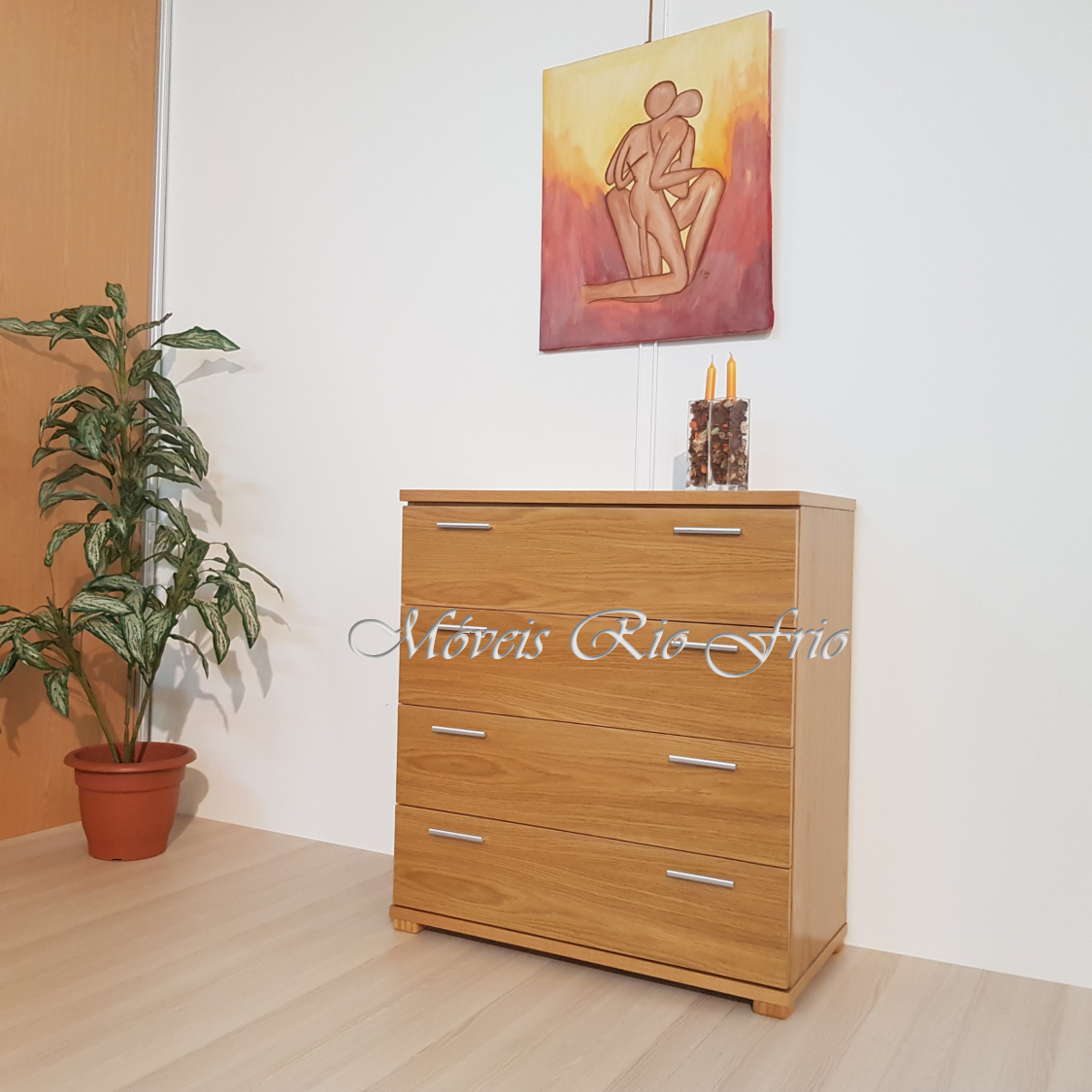 Cômoda de madeira clara com quatro gavetas, vaso decorativo com velas, quadro abstrato e planta em vaso terracota