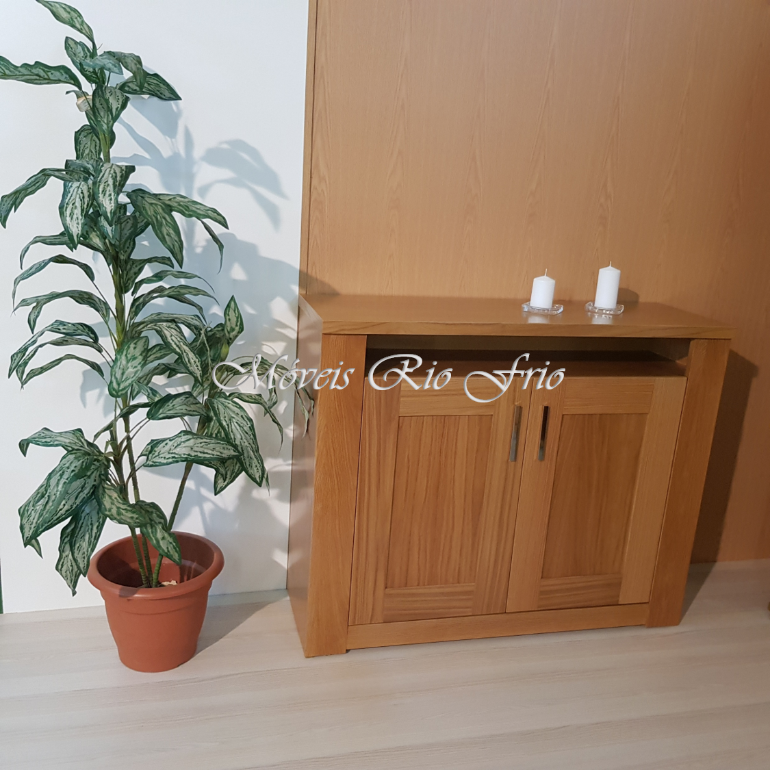 Aparador de madeira clara com duas portas e duas velas brancas em cima, planta artificial ao lado, fundo parede branca e madeira clara