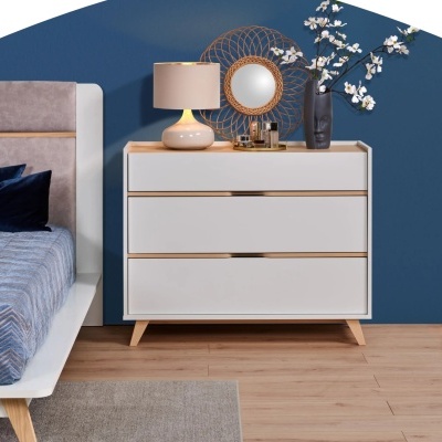 Cómoda branca com três gavetas e detalhes em madeira clara ao lado de cama e parede azul