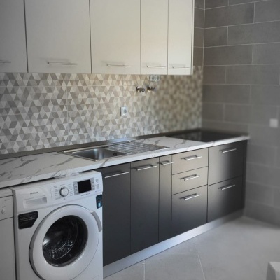 Cozinha moderna com armários brancos e cinza, bancada mármore, lava-loiça inox, máquina de lavar roupa branca e azulejos geométricos.