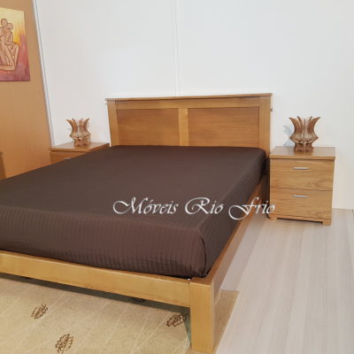 Cama de madeira clara com cabeceira, colchão com lençol preto, duas mesas de cabeceira com gavetas e lâmpadas decorativas