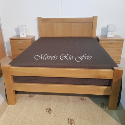 Cama de madeira clara com colcha castanha e mesas de cabeceira com lâmpadas