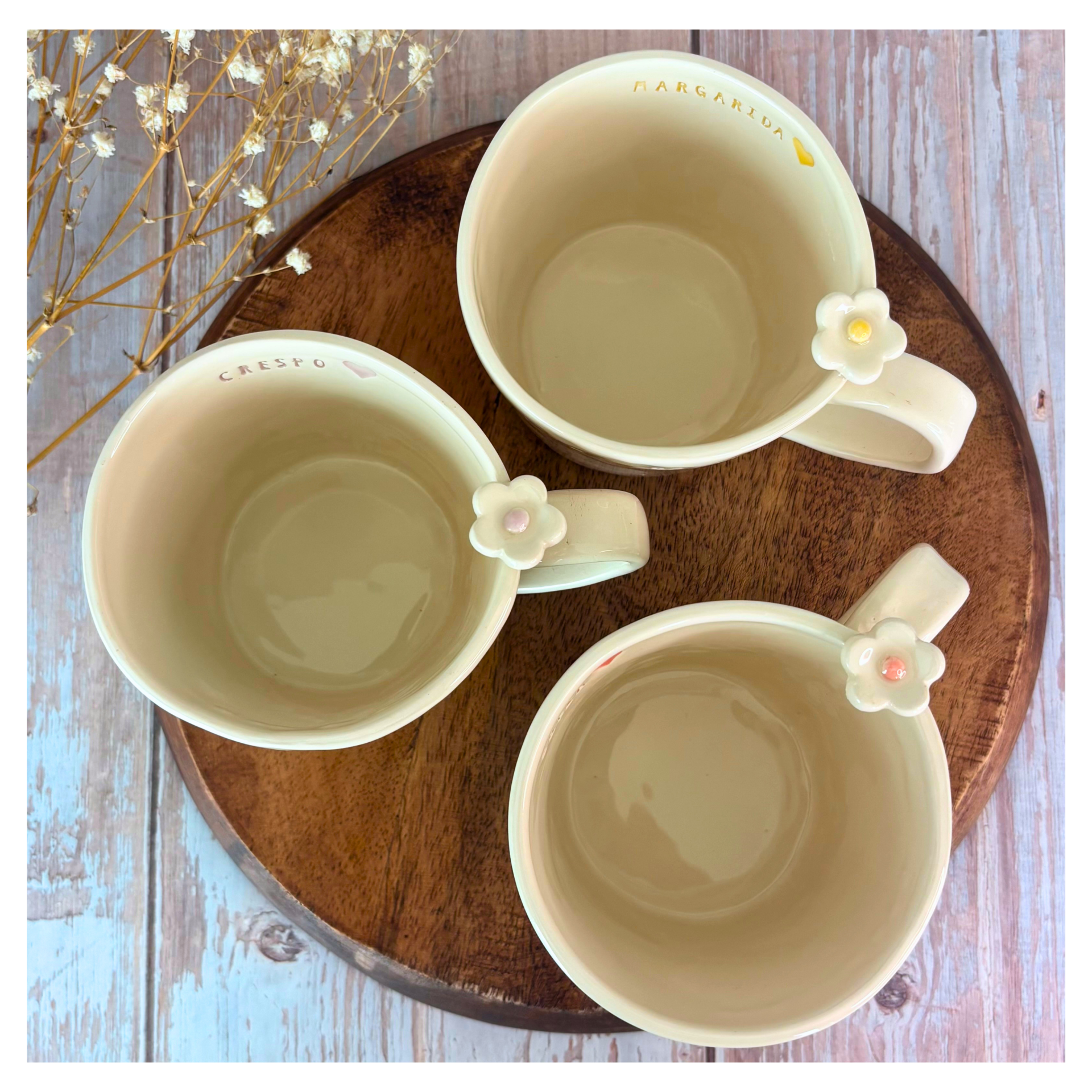 Caneca Personalizada Três canecas creme com flor na pega e texto dentro de uma delas MARGARIDA, sobre base de madeira redonda e flores secas ao lado.