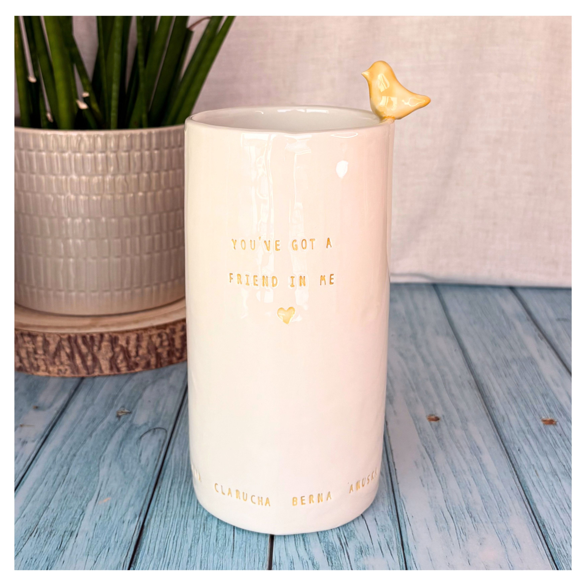 Jarra Grande Personalizada Caneca branca com pássaro amarelo e texto dourado, sobre mesa de madeira azul e planta ao fundo