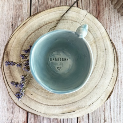 Caneca de cerâmica azul com MADRINHA gravado no interior, sobre prato de madeira e flores secas