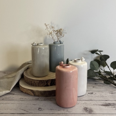 Quatro vasos cilíndricos de cerâmica com pequenos pássaros no topo, em várias cores, sobre tábuas de madeira em superfície branca envelhecida