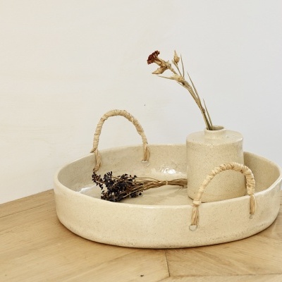 Bandeja redonda de cerâmica bege com pegas de corda e um vaso com flores secas