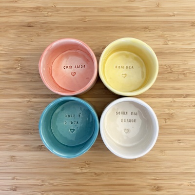 Quatro tigelas de cerâmica coloridas com frases gravadas dispostas em cima de madeira clara
