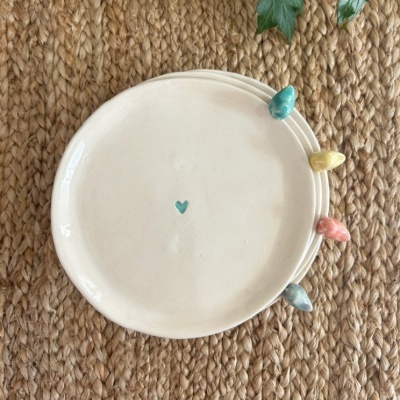 Prato de cerâmica branco com coração azul e passarinhos coloridos no bordo sobre superfície de fibra natural.