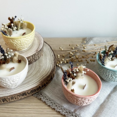 Quatro velas em recipientes cerâmicos coloridos com flores secas e pavios de madeira, sobre madeira e tecido cinza