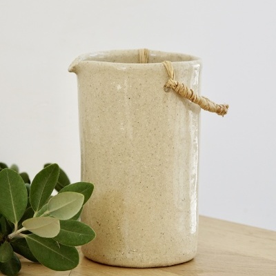 Jarro de cerâmica bege com alça de corda junto a folhas verdes sobre mesa de madeira