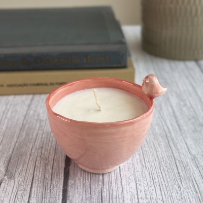 Vela em copo cerâmico rosa com pássaro decorativo sobre mesa de madeira clara
