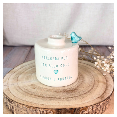 Vaso de cerâmica branco com texto azul e pássaro azul no topo sobre base de madeira com flores secas