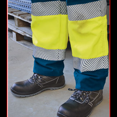 Calças de trabalho com faixas refletoras amarelas e cinzentas e sapatos pretos de segurança em ambiente industrial