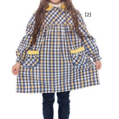 Menina com vestido de riscas quadradas azuis, brancas e amarelas, gola amarela e bolsos, calças azuis e sapatos pretos.