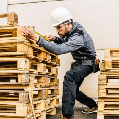 Homem a medir paletes de madeira clara com fita métrica, equipado com capacete e roupa de proteção.
