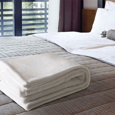 Toalha branca dobrada sobre cama com colcha cinza texturada e almofadas brancas num quarto