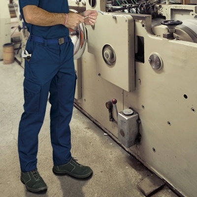 Homem com calças de trabalho azuis e sapatos de segurança verdes junto a máquina industrial bege