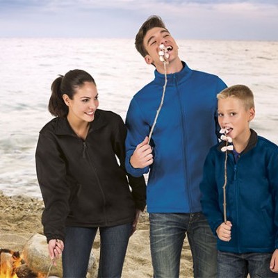 Três pessoas de casacos fleece seguram pauzinhos com marshmallows junto a fogueira na praia.