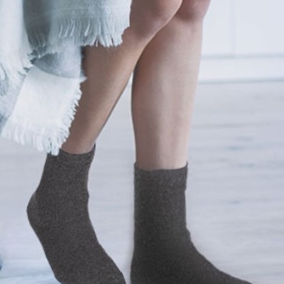 Meias castanho-escuro com textura suave usadas numa pessoa de pé em chão de madeira claro, com cobertor cinza e branco ao lado