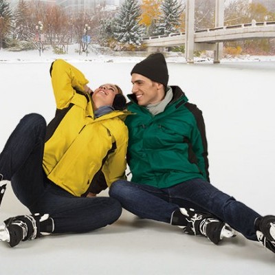 Casal sentado no gelo com patins, vestindo casacos amarelo e verde