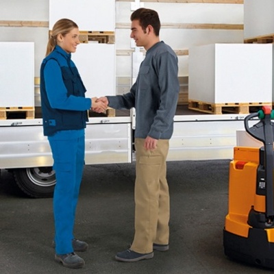 Dois trabalhadores cumprimentando-se numa área de armazenamento industrial com empilhadora elétrica amarela e caixas em paletes