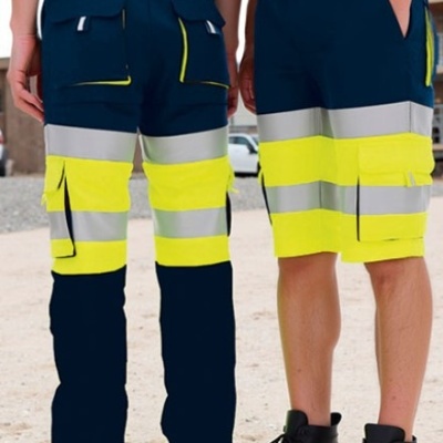 Calças e calções de trabalho alta visibilidade azul escuro com faixas amarelas e refletoras, com botas pretas, em chão exterior arenoso