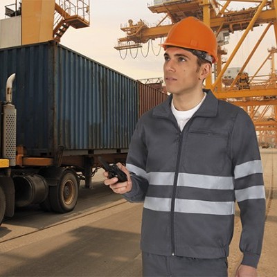Homem com uniforme cinzento e capacete laranja num cais industrial com camião e contentor