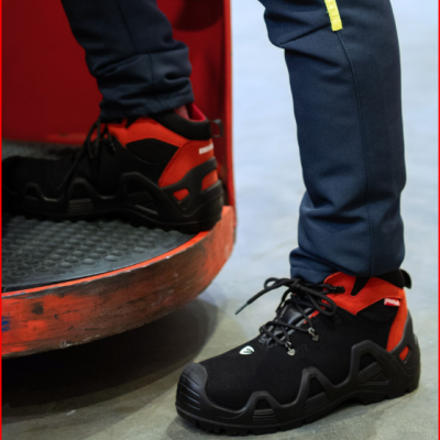 Tênis desportivos preto e vermelho com sola preta robusta usados com calças azuis junto a piso cinzento e elemento vermelho