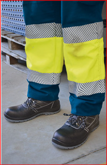 Calças de trabalho com faixas refletoras amarelas e cinzentas e sapatos pretos de segurança em ambiente industrial