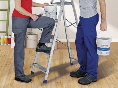 Duas pessoas com roupas de trabalho, uma na escada e outra de pé, cada uma com um balde de tinta branca.