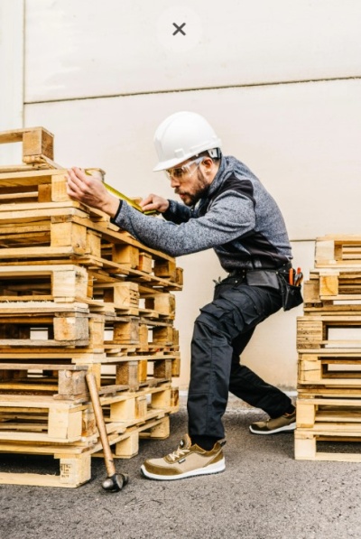 Homem a medir paletes de madeira clara com fita métrica, equipado com capacete e roupa de proteção.