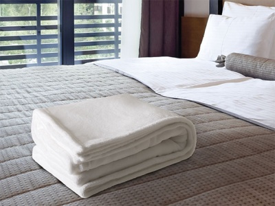 Toalha branca dobrada sobre cama com colcha cinza texturada e almofadas brancas num quarto